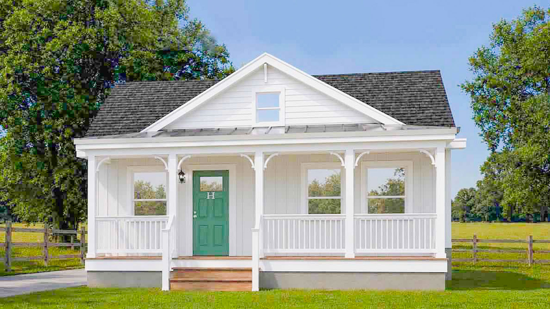Small white modular home with a green door, front porch, and steps, set in a grassy yard with trees and a fence.